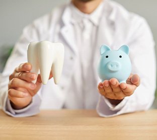 Dentist in white coat holding model tooth and blue piggy bank